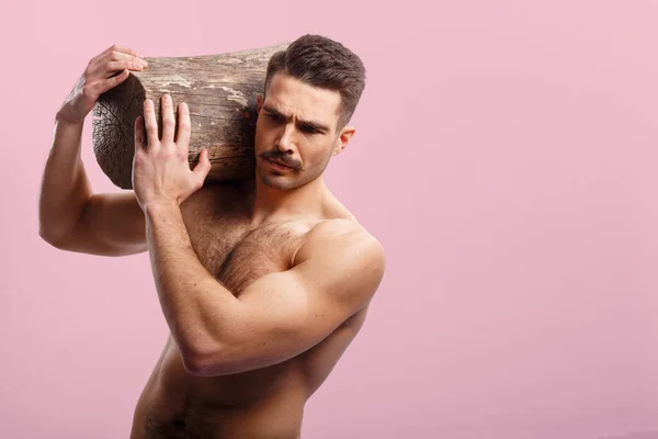 背景にピンクの上に彼の肩に木の幹を保持し 裸の胴を持つアスリート セクシー 残忍なひげを生やした男 — ストック写真