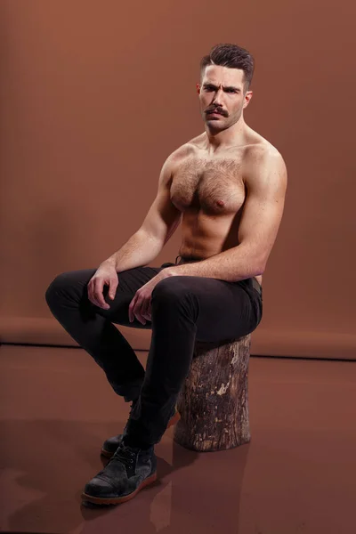 残酷なひげを生やした男は 恥知らずで 木の幹 茶色の背景に座って筋肉質の体 二足歩行やトリップを示しています 垂直表示 — ストック写真