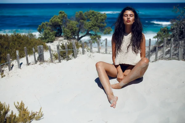 Une belle jeune fille aux longs cheveux bruns, assise sur le sable chaud aux yeux fermés, sur fond de plage de Corse. — Photo