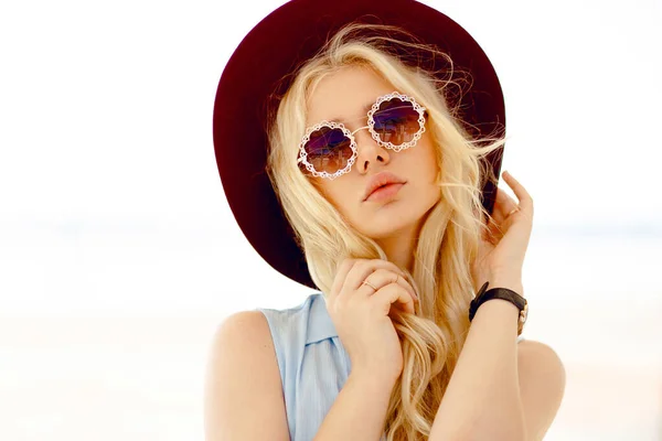 Sensual chica rubia con gafas de sol redondas florales, pelo rizado, labios grandes y sombrero toca su propio cabello y mirando a la cámara. — Foto de Stock