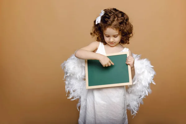 Adorável anjo pequena menina encaracolado com asas de anjo segurando e apontando o dedo em uma placa, sobre fundo bege. — Fotografia de Stock