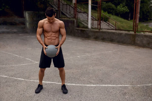 Подходит сексуальный мускулистый молодой человек, стоящий на баскетбольной площадке и держащий серый мяч в руках, на заднем плане улицы. — стоковое фото