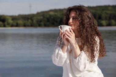 Sahildeki bir parkta duran kahve fincanı kokan tatmin olmuş genç bir kadının yan görüntüsü..