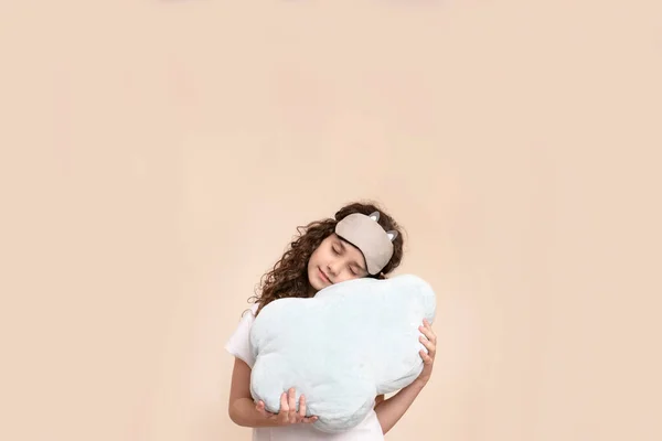 Little girl with curly hair isolated on a beige background, holding a pillow, have a sleeping mask. — Stock Photo, Image