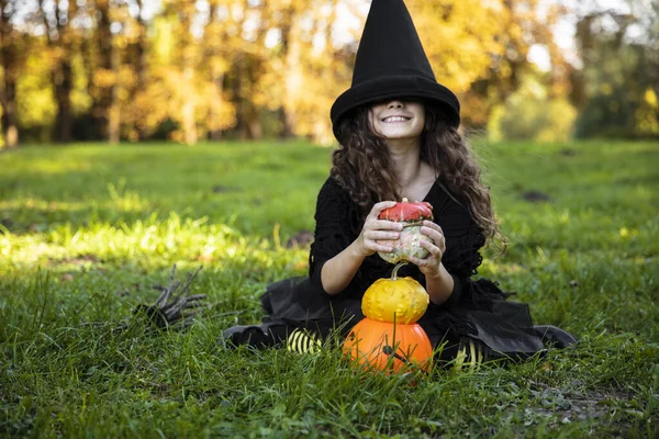 Adorable petite fille aux cheveux en chapeau de sorcière noir faisant grimace, tenant des citrouilles, sur un fond ensoleillé d'automne. — Photo