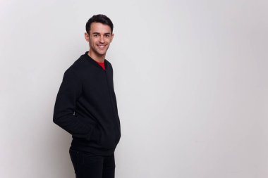 Koyu mavi kazaklı ve kot pantolonlu esmer adam beyaz duvar arkasında poz veriyor..