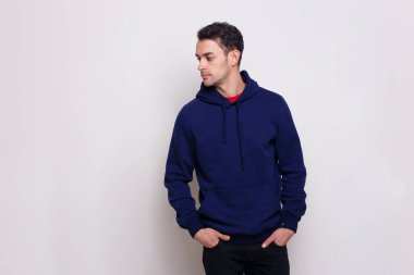 Koyu mavi kazaklı ve kot pantolonlu bir gencin profili. Ellerini cebinde tutuyor. Gri bir arka planı var..