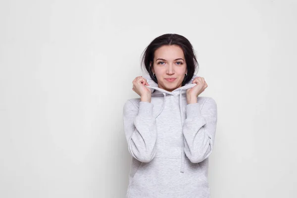 Giovane donna bruna in un maglione grigio chiaro con cappuccio, guardando la fotocamera isolata su sfondo bianco. Copia spazio. — Foto Stock