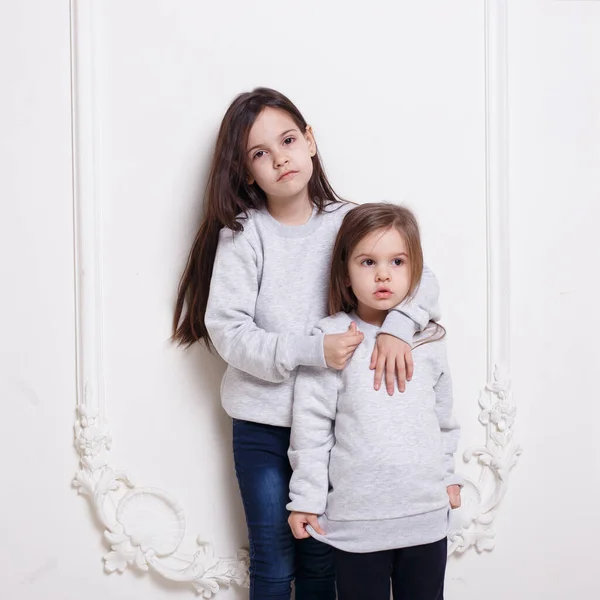 Дві чарівні маленькі дівчинки в светрі і джинсах, позують разом, обійняті на білому тлі . — стокове фото