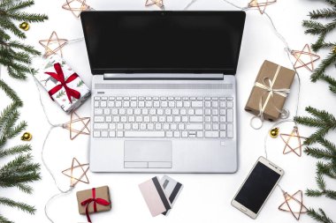 Noel alışverişi. Beyaz arka planda açık bir laptop, bir kredi kartı, kırmızı kurdeleli hediye kutuları, yeşil köknar dalları, yıldız çelenkleri. Kış tatilinde online ticaret.