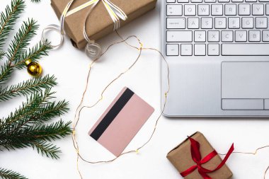 Noel alışverişi. Beyaz arka planda açık bir laptop, bir kredi kartı, kırmızı kurdeleli hediye kutuları, yeşil köknar dalları, yıldız çelenkleri. Kış tatilinde online ticaret.