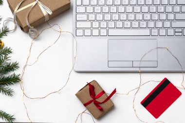 Noel alışverişi. Beyaz arka planda açık bir laptop, bir kredi kartı, kırmızı kurdeleli hediye kutuları, yeşil köknar dalları, yıldız çelenkleri. Kış tatilinde online ticaret.