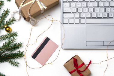Noel alışverişi. Beyaz arka planda açık bir laptop, bir kredi kartı, kırmızı kurdeleli hediye kutuları, yeşil köknar dalları, yıldız çelenkleri. Kış tatilinde online ticaret.