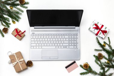 Noel alışverişi. Beyaz arka planda açık bir laptop, bir kredi kartı, kırmızı kurdeleli hediye kutuları, yeşil köknar dalları, yıldız çelenkleri. Kış tatilinde online ticaret.