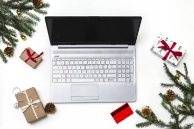 Noel alışverişi. Beyaz arka planda açık bir laptop, bir kredi kartı, kırmızı kurdeleli hediye kutuları, yeşil köknar dalları, yıldız çelenkleri. Kış tatilinde online ticaret.