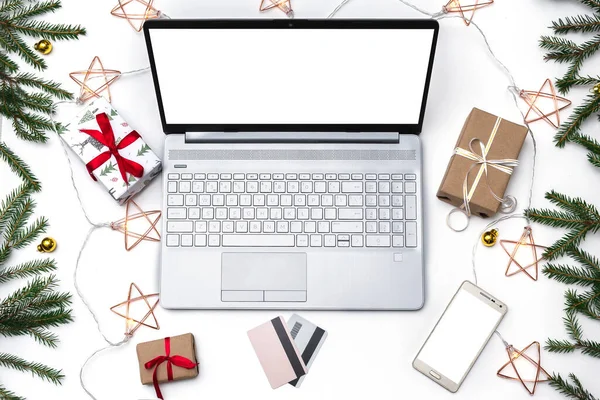 Noel alışverişi. Beyaz arka planda açık bir laptop, bir kredi kartı, kırmızı kurdeleli hediye kutuları, yeşil köknar dalları, yıldız çelenkleri. Kış tatilinde online ticaret.