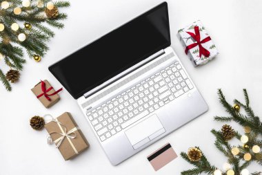 Noel alışverişi. Beyaz arka planda açık bir laptop, bir kredi kartı, kırmızı kurdeleli hediye kutuları, yeşil köknar dalları, yıldız çelenkleri. Kış tatilinde online ticaret.