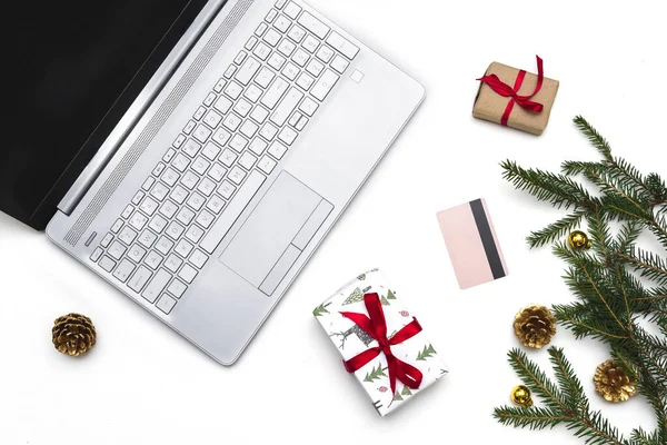 Noel alışverişi. Beyaz arka planda açık bir laptop, bir kredi kartı, kırmızı kurdeleli hediye kutuları, yeşil köknar dalları, yıldız çelenkleri. Kış tatilinde online ticaret.