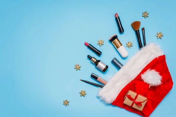 Makyaj ürünleri ve mavi arka planda Noel dekoru olan düz yatak. Makyaj fırçaları, rujlar ve Noel Baba 'nın kırmızı şapkasından çıkan bir kavanoz yağ. Üstten manzara. Kadınların dilekleri kavramı.