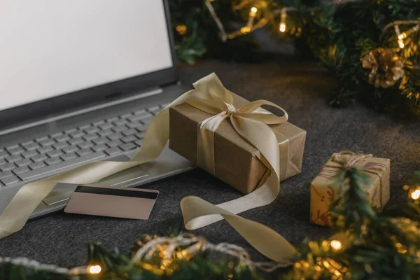 Noel alışverişi için kredi kartıyla internetten. Masanın üzerinde hediyeler olan dizüstü bilgisayar köknar dalları ve ışık çelengi..