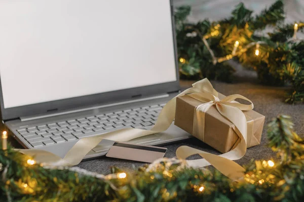 Noel alışverişi için kredi kartıyla internetten. Masanın üzerinde hediyeler olan dizüstü bilgisayar köknar dalları ve ışık çelengi..