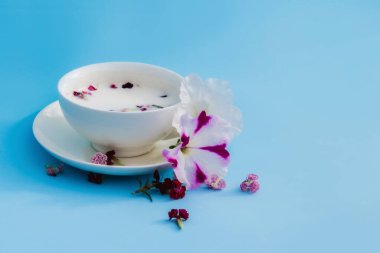 Mavi arka planda vejetaryen sütü ve çiçekli beyaz kupa. Vejetaryen içecek ve yiyecek kavramı, bitki çayı, güzellik ve sağlık, spa.