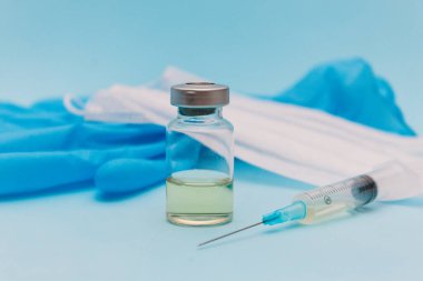 Aşı bir kavanozda, bir şırınganın yanında, tıbbi bir maske ve mavi bir arka planda mavi eldivenler..