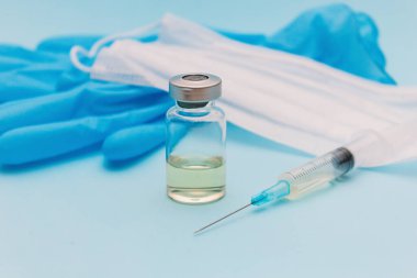 Aşı bir kavanozda, bir şırınganın yanında, tıbbi bir maske ve mavi bir arka planda mavi eldivenler..