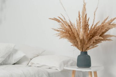 İskandinav ve Boho tarzında beyaz minimalist yatak odası. Komodinin üstündeki vazoda bir bukette otlar..