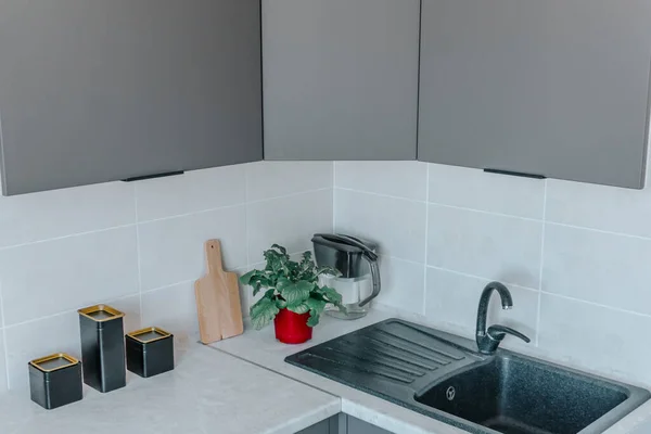 On the kitchen countertop are a chopping board, black containers, and a flower in a red board, next to the sink. The concept of minimalism and environmental friendliness in the kitchen.