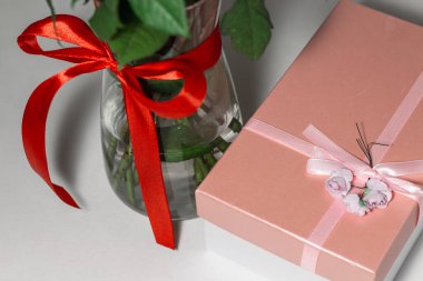 Beyaz çay gülleri kırmızı kurdeleyle bağlanmış ve masanın üzerinde duran cam bir vazoda duruyor. Yakınlarda bir hediye var. Pembe bir kutuda. Hediye buketi.