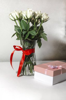 Beyaz çay gülleri kırmızı kurdeleyle bağlanmış ve masanın üzerinde duran cam bir vazoda duruyor. Yakınlarda bir hediye var. Pembe bir kutuda. Hediye buketi.