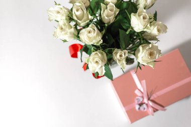 Beyaz çay gülleri kırmızı kurdeleyle bağlanmış ve masanın üzerinde duran cam bir vazoda duruyor. Yakınlarda bir hediye var. Pembe bir kutuda. Hediye buketi.