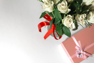 Beyaz çay gülleri kırmızı kurdeleyle bağlanmış ve masanın üzerinde duran cam bir vazoda duruyor. Yakınlarda bir hediye var. Pembe bir kutuda. Hediye buketi.