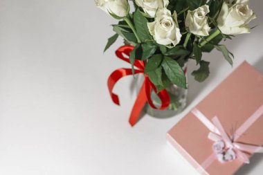 Beyaz çay gülleri kırmızı kurdeleyle bağlanmış ve masanın üzerinde duran cam bir vazoda duruyor. Yakınlarda bir hediye var. Pembe bir kutuda. Hediye buketi.