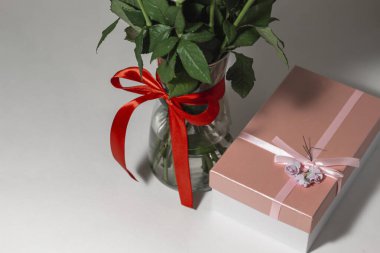 Beyaz çay gülleri kırmızı kurdeleyle bağlanmış ve masanın üzerinde duran cam bir vazoda duruyor. Yakınlarda bir hediye var. Pembe bir kutuda. Hediye buketi.