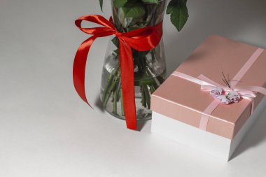 Beyaz çay gülleri kırmızı kurdeleyle bağlanmış ve masanın üzerinde duran cam bir vazoda duruyor. Yakınlarda bir hediye var. Pembe bir kutuda. Hediye buketi.