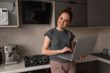 Neşeli, çekici bir kız, gri bir mutfakta dizüstü bilgisayarla çalışırken gülümsüyor..