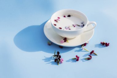 Mavi arka planda vejetaryen sütü ve çiçekli beyaz kupa. Vejetaryen içecek ve yiyecek kavramı, bitki çayı, güzellik ve sağlık, spa.