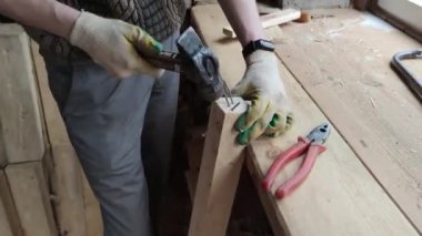 A man wearing gloves bangs a hammer on nails stuck in a wooden bar to get it out
