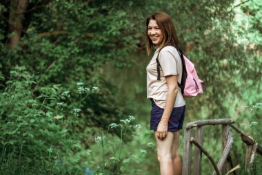 Kısa saçlı ve pembe sırt çantalı genç bir kadın ormandaki bir nehrin kenarındaki küçük tahta bir köprüde duruyor kameraya bakıyor ve gülümsüyor. seçici odak.