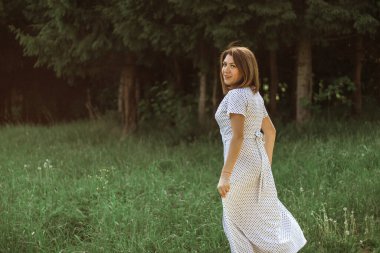 Beyaz benekli elbiseli, kısa saçlı mutlu bir genç kadın ormanın yakınındaki bir çayırda yürüyor. Doğal güzelliği olan bir kızın portresi. Seçici Odaklanma.