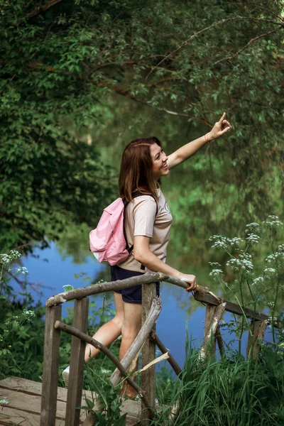 Kısa saçlı ve pembe sırt çantalı mutlu genç bir kadın ormanın yakınındaki küçük tahta bir köprünün üzerinde duruyor, gülümsüyor ve uzaklığı işaret ediyor. Seçici Odaklanma.
