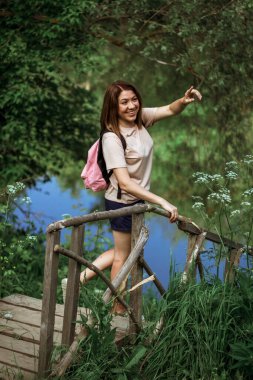 Kısa saçlı ve pembe sırt çantalı mutlu genç bir kadın ormanın yakınındaki küçük tahta bir köprünün üzerinde duruyor, gülümsüyor ve uzaklığı işaret ediyor. Seçici Odaklanma.