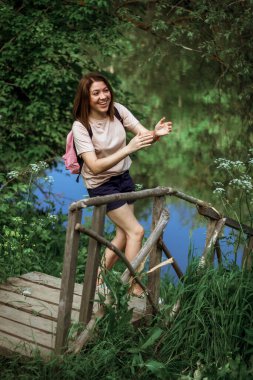 Kısa saçlı ve pembe sırt çantalı mutlu genç bir kadın ormanda bir nehrin kenarındaki küçük tahta bir köprüde duruyor, gülümsüyor ve ellerini çırpıyor. Seçici Odaklanma.