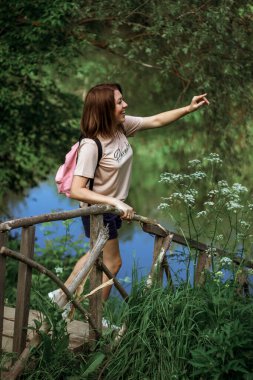 Kısa saçlı ve pembe sırt çantalı mutlu genç bir kadın ormanın yakınındaki küçük tahta bir köprünün üzerinde duruyor, gülümsüyor ve uzaklığı işaret ediyor. Seçici Odaklanma.