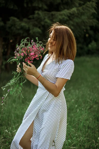 白いドレスの美しい幸せな女の子は彼女の手と笑顔で野生の花の花束を保持しています 森の近くの牧草地で野生の花の花束と陽気な女の子の肖像画 選択的焦点 — ストック写真