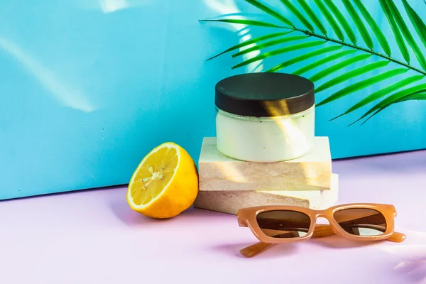Cosmetics are on the podium on a summer background, in the rays of the sun. a jar of cream is on the podium, half a lemon and sunglasses are lying next to it, and a palm leaf is hanging above it.
