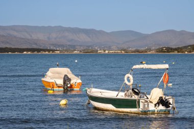 İyon Denizi koyunda iki tekne. Orman ve binalarla dolu bir sahil. Arka planda dağlar. Mavi yaz gökyüzü. Roda, Korfu (Korfu), Yunanistan.