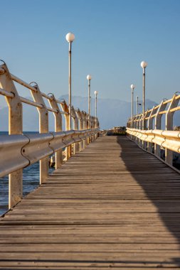 Yıpranmış ahşap zemini ve paslı parmaklıkları olan eski iskele. İki tarafta da sokak lambaları var. İyon denizi ve mavi gökyüzü. Roda, Korfu (Korfu), Yunanistan.